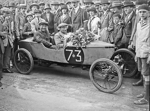 A small pointed racing car numbered 73 with a bouquet on its scuttle, two men aboard, a dense crowd in hats around it. Opens a larger view