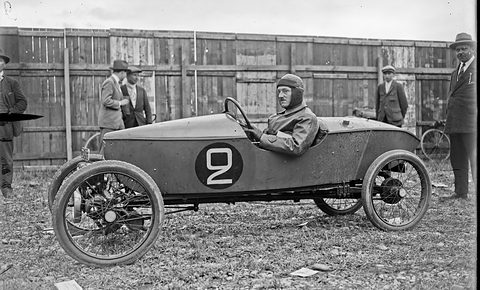 Side view of a long low pointed cyclecar with wire wheels and a large number 2, its helmeted driver seated, men behind a fence. Opens a larger view