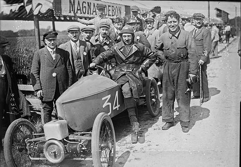 A helmeted driver in a leather coat in a small pointed cyclecar numbered 34, a grinning mechanic in overalls and officials round him. Opens a larger view