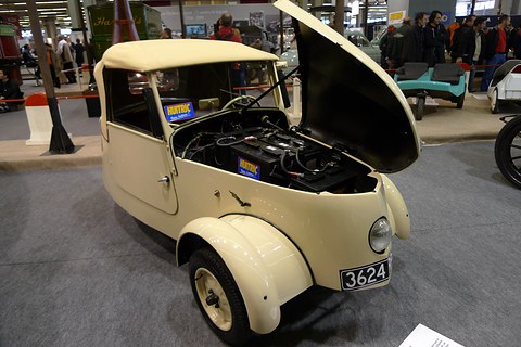 Cream two-seater with its one-piece bonnet tipped up and batteries visible inside, show visitors in the background.