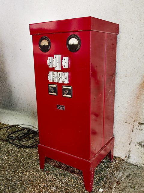 Tall red metal cabinet with two round meters and switch panels, standing on gravel against a white wall. Opens a larger view