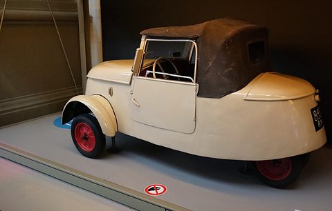 Cream two-seater with a brown hood and red wheels standing on a raised plinth inside a glass case. Opens a larger view