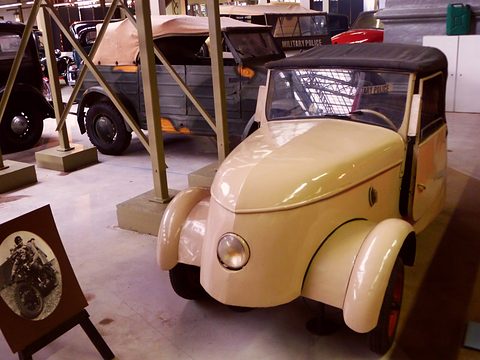 Cream two-seater with a brown hood seen close from the right front, wooden-framed exhibits and old cars behind. Opens a larger view
