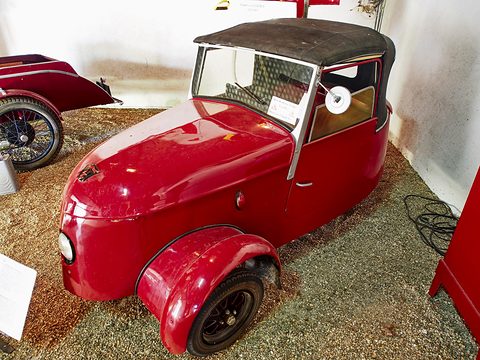 Red two-seater with a black fabric hood and single headlamp, standing on gravel in a whitewashed museum hall. Opens a larger view