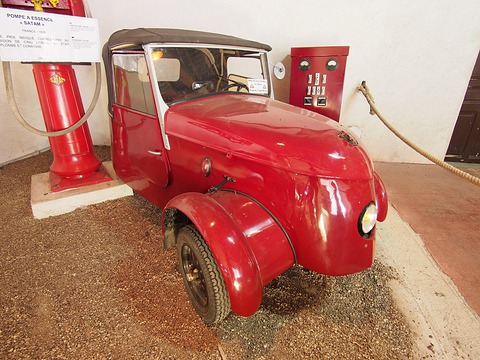 Red electric two-seater angled towards the camera, a red petrol pump and a red electrical cabinet with meters behind it. Opens a larger view