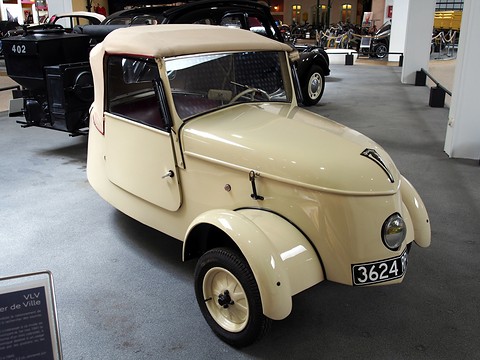 Small cream two-seater with a single round headlamp in its rounded nose, cycle-type front wings and a beige fabric hood, on a museum floor. Opens a larger view