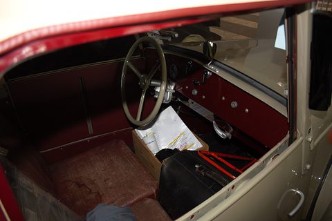 View through the open door: large steering wheel, plain painted dashboard, worn red floor and papers on the seat. Opens a larger view
