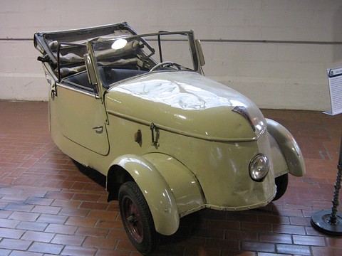 Scuffed pale green two-seater with its hood folded back and a dented nose, on a brick floor against a white wall. Opens a larger view