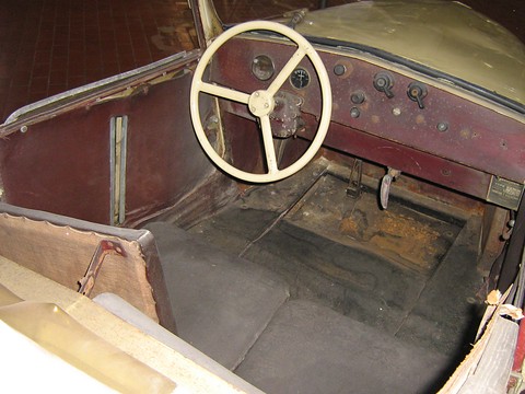 Cream three-spoke steering wheel, a small dashboard with instruments and a bare, rusted floor between the seats. Opens a larger view