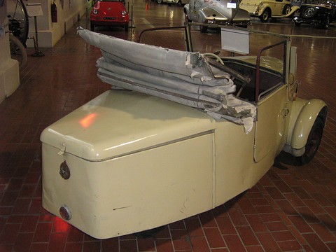 Rear three-quarter of a worn pale car with its folded hood piled on the tail and closely set rear wheels. Opens a larger view
