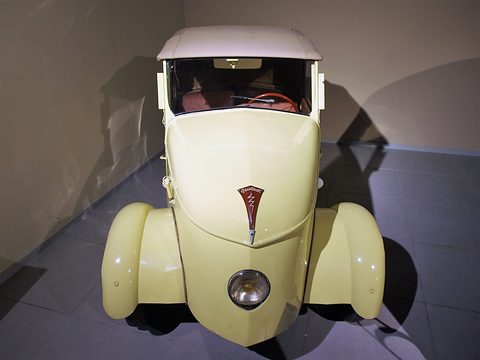 Pale yellow narrow car from straight ahead, a single headlamp under the lion badge, white hood, dark display floor. Opens a larger view