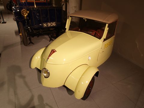 Pale yellow two-seater lettered POSTES on its door, seen from above the left front, a veteran car behind.