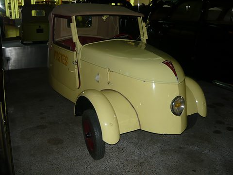 Pale yellow two-seater lettered POSTES, close from the right front, lit by flash against a dark background. Opens a larger view