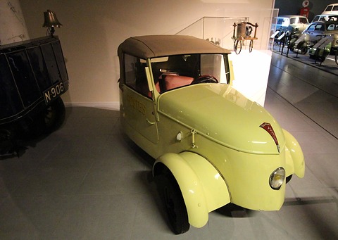 Yellow two-seater with its grey hood up, seen from the right front in a dark museum hall. Opens a larger view