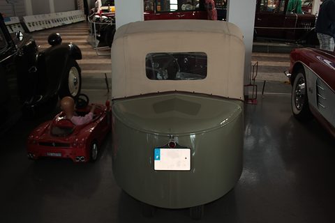 Grey-green car from directly behind: tall fabric hood with a small window and a rounded tail, a child’s pedal car beside it. Opens a larger view