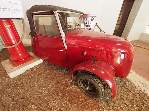 Red two-seater from the right with its black hood up, a tall red petrol pump on the left and a rope barrier behind. Opens a larger view