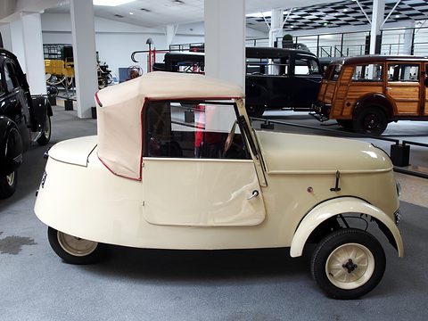 Cream two-seater from the right side, showing its short body, a single door, the raised fabric hood and the rounded tail. Opens a larger view
