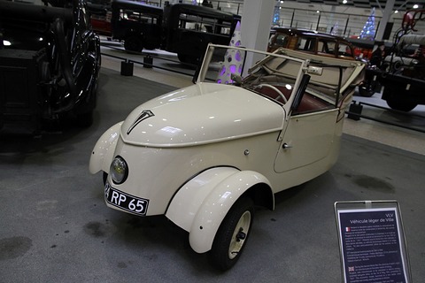 Cream open two-seater with its windscreen frame up and the hood stowed, a museum information panel beside it. Opens a larger view
