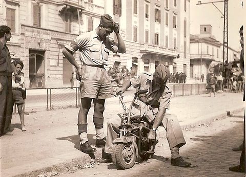 Two soldiers in shorts and berets, one crouching astride a tiny folding motorcycle on a city pavement, onlookers behind.