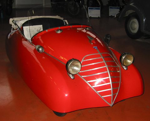 Head-on view of a small red open car with a pointed chrome-barred grille and two round headlamps, in a dark hall.
