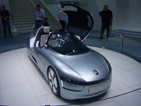 Silver low tandem two-seater with its canopy lifted on a white turntable, visitors standing behind. Opens a larger view