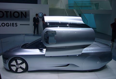 Side view of a silver, low tandem-seat streamliner with a long canopy lifted, a VW sign behind. Opens a larger view