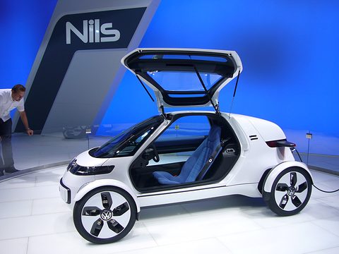 White single-seat concept with a gullwing door raised against a bright blue Nils backdrop. Opens a larger view