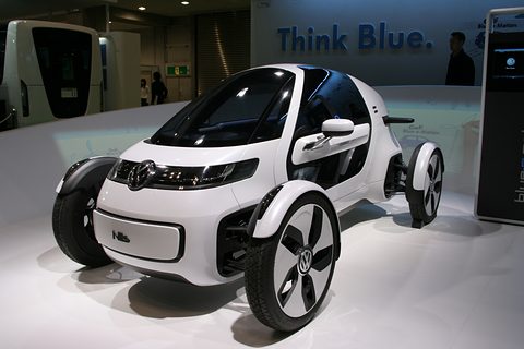 White single-seat concept with open wheels and a black canopy on a white stand under a ‘Think Blue.’ sign. Opens a larger view