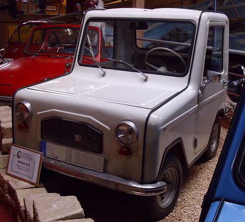 Pale grey boxy two-seat microcar with a black grille panel and round headlamps, parked on gravel among other small cars. Opens a larger view