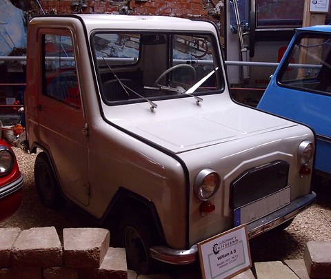 Pale grey boxy microcar seen from the front right, its windscreen wipers raised, a blue car and a red car beside it. Opens a larger view