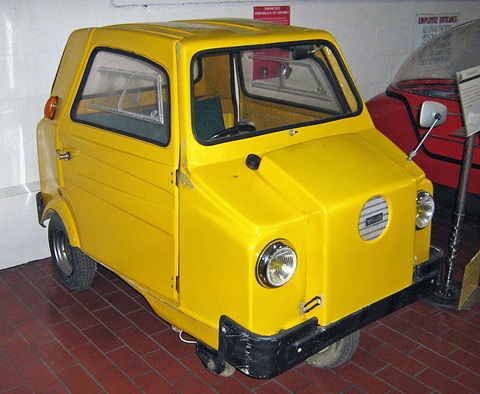 Yellow single-seat three-wheeler with a box cowl at the nose, a round grille and black bumper, on a red-tiled floor. Opens a larger view