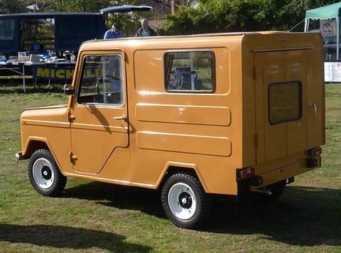 Ochre boxy van seen from the rear right, its single rear door with a small window, show stalls and gazebos behind. Opens a larger view