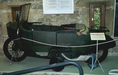 A long dark green narrow car with a folding windscreen and spoked wheels behind a rope, an information placard beside it.