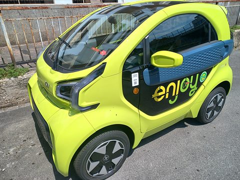 Lime-green two-seat microcar with a blue door panel and ‘enjoy’ lettering parked by a railing. Opens a larger view