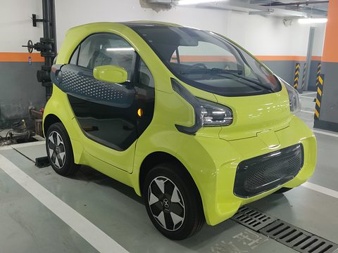 Lime-green and black two-seat microcar with slim LED lamps and a mesh grille in a car park. Opens a larger view