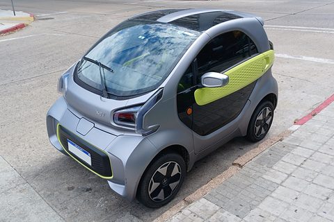 Silver two-seat microcar with lime-green door trim parked at a kerb beside a tiled pavement. Opens a larger view