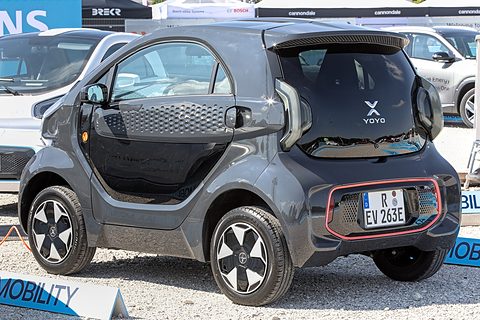 Rear of a grey two-seat microcar with a black glass tailgate, X badge and red-trimmed bumper on gravel. Opens a larger view