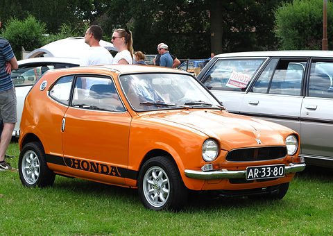 Orange small coupé with black HONDA lettering on the sill, parked on grass with visitors behind. Opens a larger view