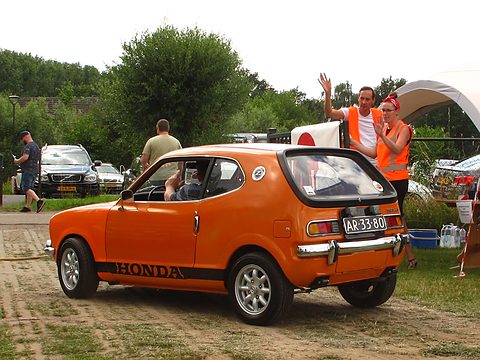 Rear three-quarter of an orange hatchback with HONDA on its sill, marshals waving behind. Opens a larger view