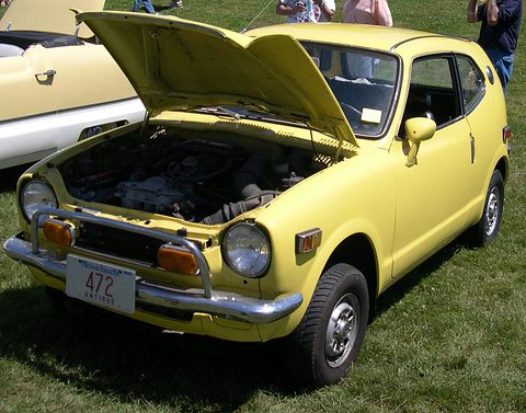 Yellow small hatchback on grass with the bonnet raised over its engine, another car at left. Opens a larger view