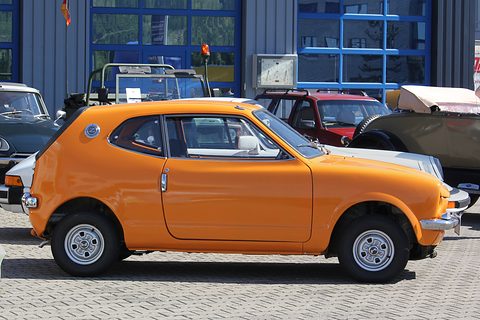 Side view of an orange small coupé on paving in front of a blue building. Opens a larger view