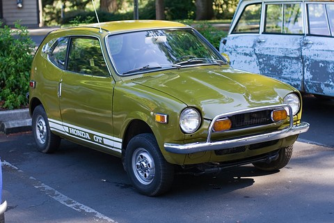Olive-green small coupé with white lettered stripes along the sill, in a car park. Opens a larger view