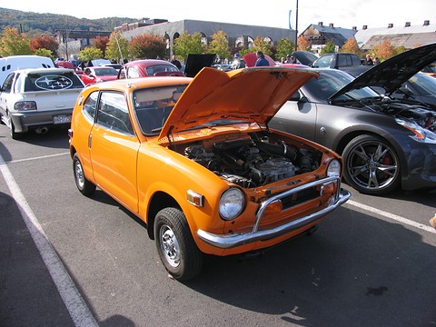 Orange small coupé with the bonnet up in a busy car park full of show cars. Opens a larger view