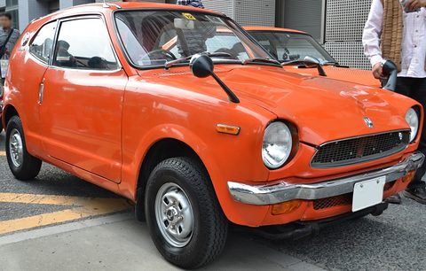 Orange small coupé with round headlamps, a man in a white shirt standing at its right. Opens a larger view