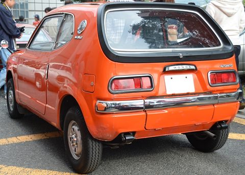 Rear view of an orange hatchback with a black-framed rear window, visitors at left. Opens a larger view