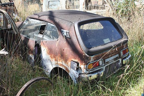 A rusty, faded small hatchback among long grass and scrap in a wrecking yard. Opens a larger view