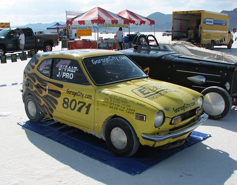 Yellow small coupé covered in sponsor decals and racing number 8071 on a blue mat on white salt. Opens a larger view
