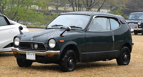 Black small coupé with round headlamps and raindrops on its paint, parked on dry grass. Opens a larger view
