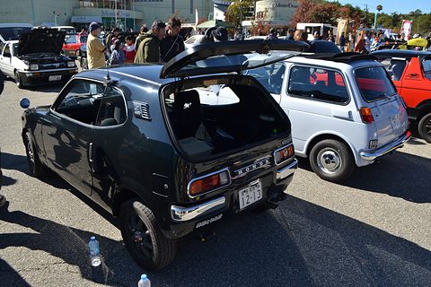 Black hatchback with the rear glass lifted, a silver small car beside it and a crowd behind. Opens a larger view