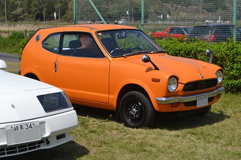 Orange small coupé parked on grass in front of a hedge, a white car at left. Opens a larger view